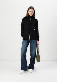 Maglione nero con colletto alto e bottoni davanti, abbinato a jeans in denim blu a campana e sneakers bianche. La modella tiene una borsa verde oliva.