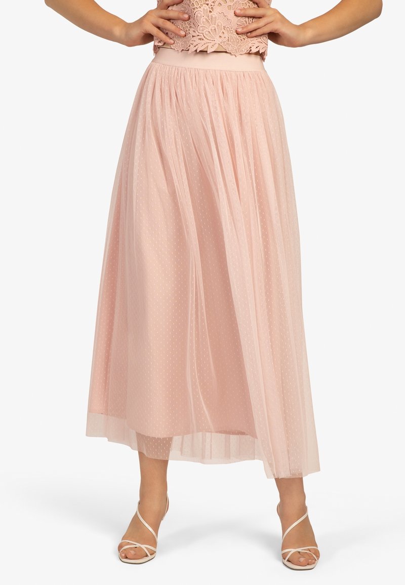 Femme portant une jupe midi taille haute rose pâle avec un overlay en tulle à pois et des sandales blanches à talons ouverts sur fond blanc.