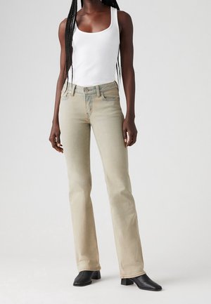 Kvinde iført en hvid tanktop, lysebeige jeans med lige ben og sorte hælede støvler med firkantet tå, stående mod en ensfarvet baggrund.