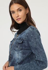 Chaqueta de denim en azul con un lavado deslavado, cierre de cremallera, dos bolsillos frontales y un corte estructurado. Presenta mangas largas y un diseño de cuello clásico.