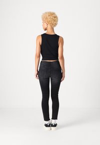 Fekete cropped tank top, illeszkedő fekete farmernadrág enyhe kopással, és fekete-fehér sportcipő. A modellt hátulról nézzük, egyszínű háttér előtt.