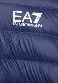 Marineblå pufferjakke med horisontal søm og en blank overflate. Har en fremtredende hvit EA7 Emporio Armani-logo.
