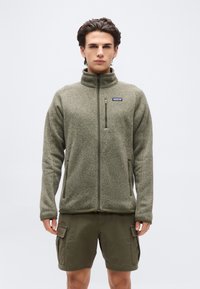 Άνδρας φορώντας ζακέτα φλις με πλήρες φερμουάρ σε ελιά πράσινο χρώμα με λογότυπο στο στήθος και ταιριαστό cargo σορτς, που στέκεται μπροστά σε απλό φωτεινό φόντο.