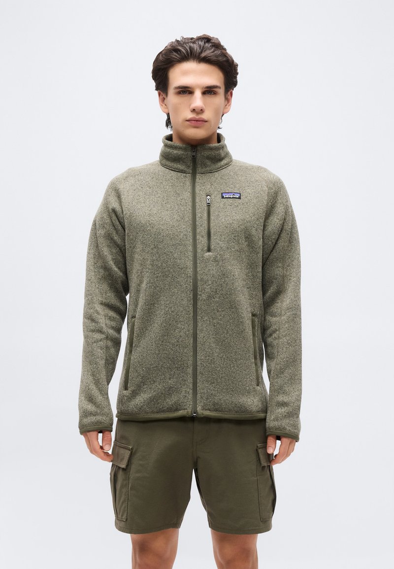 Άνδρας φορώντας ζακέτα φλις με πλήρες φερμουάρ σε ελιά πράσινο χρώμα με λογότυπο στο στήθος και ταιριαστό cargo σορτς, που στέκεται μπροστά σε απλό φωτεινό φόντο.