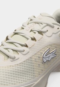 Close-up van een beige Lacoste sneaker met witte veters, een mesh-textuur en een geborduurd krokodillenlogo aan de zijkant.
