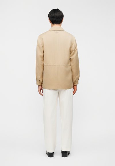 Μπεζ σακάκι με γιακά, μανσέτες με κουμπιά και ίσια γραμμή, σε συνδυασμό με λευκό παντελόνι και μαύρα παπούτσια, που φαίνεται από πίσω.