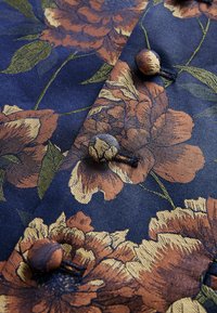 Marineblauer Stoff mit einem großen Blumenmuster in orange und goldenen Farbtönen. Vier dekorative Knöpfe in Braun und Gold, die die Textur des Designs betonen.