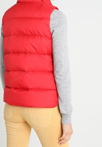 Rode puffer-vest met horizontale quiltlijnen, hoge hals en zijzakken. Draag het over een grijs longsleeve shirt, gecombineerd met lichtgekleurde broek.
