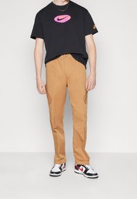 Svart t-shirt med en rosa och lila Nike-logotyp, kombinerad med sandfärgade cargo-byxor med sidfickor och svartvita sneakers.