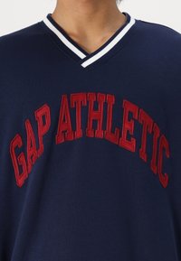 Granatowy bawełniany t-shirt z dekoltem w serek i białym paskiem oraz dużym czerwonym haftowanym napisem „GAP ATHLETIC” na piersi.