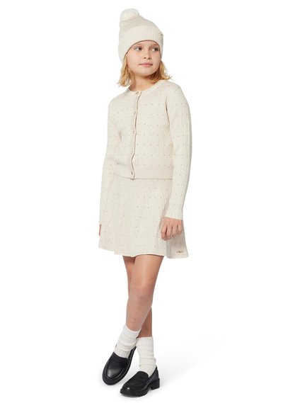 Strikket cardigan og skjørtsett i krem, med teksturerte mønstre. Kombinert med en matchende lue og svarte loafers, med hvite sokker.