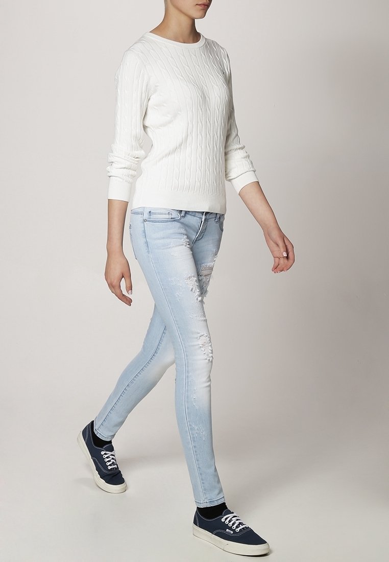Witte kabelgebreide sweater, lichtblauwe versleten skinny jeans en marineblauwe sneakers met witte veters. Model loopt tegen een neutrale achtergrond.