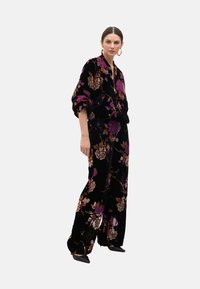 Černý sametový outfit s květinovými vzory v purpurové a hnědé barvě, s volnou blůzou a kalhotami širokého střihu, doplněný špičatými černými botami.
