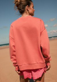 Koralle Sweatshirt mit überschnittenen Schultern und gerippten Bündchen, kombiniert mit rosa Karo-Shorts. Glatte Textur, lässige Passform, Strandambiente.