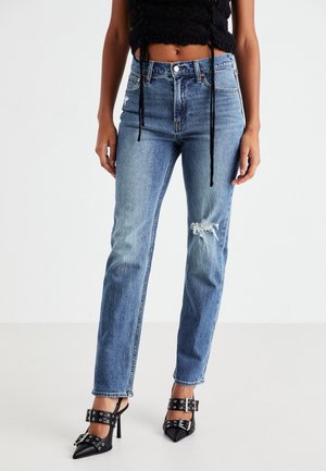 Femme portant un jean bleu taille haute avec une déchirure sur la cuisse droite, un crop top noir texturé et des talons hauts noirs pointus avec des boucles.