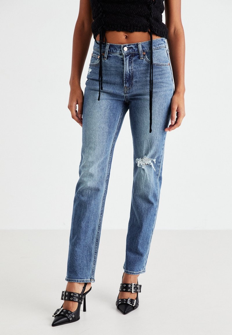 Femme portant un jean bleu taille haute avec une déchirure sur la cuisse droite, un crop top noir texturé et des talons hauts noirs pointus avec des boucles.