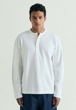 Hombre de pie de frente, lleva una camiseta Henley blanca de manga larga con tres botones y pantalones azul oscuro, sobre un fondo claro y liso.