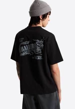 Person iført en grå hue og en sort Vans T-shirt med en stor sølvdrage og teksten "Balliro" på bagsiden.
