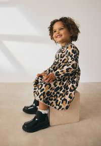 Leopardprint kjole med lange ærmer, plisseret nederdel og bølget kant; skinnende sorte ankelstøvler med hvidt blondertrim hviler på en beige blok.