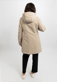 Giacca cappuccio beige con texture trapuntata, taglio lunghezza ginocchio e orlo foderato in pelliccia sintetica. Abbinata a pantaloni neri e sneakers bianche.