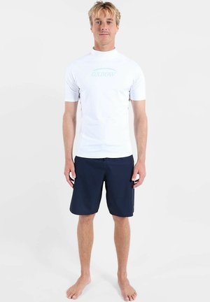 Homme blond debout pieds nus portant un rash guard blanc à manches courtes avec le logo "Oxbow" bleu clair et un short de bain bleu marine.