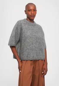 Personne chauve portant un pull court en mohair gris ample à manches courtes et un pantalon plissé marron taille haute, se tenant devant un fond blanc.
