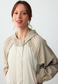 Beige leichte Jacke mit Kapuze, ausgestattet mit elastischen Bündchen, einem Reißverschluss vorne und einer glatten Textur. Entworfen mit einem entspannten Schnitt und einem dezenten Glanz.