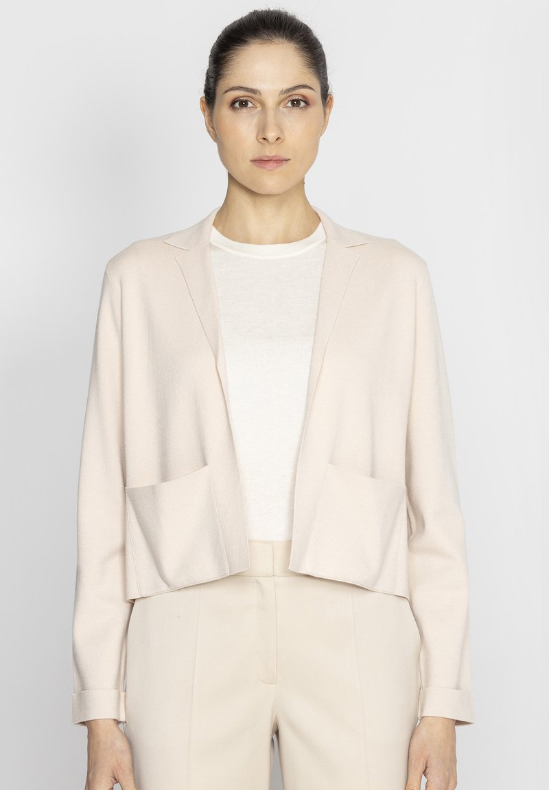 Jasnoróżowa krótkia marynarka z długimi rękawami, dwoma przednimi kieszeniami i wykładanym kołnierzem, noszona na białej bluzce i beżowych spodniach.