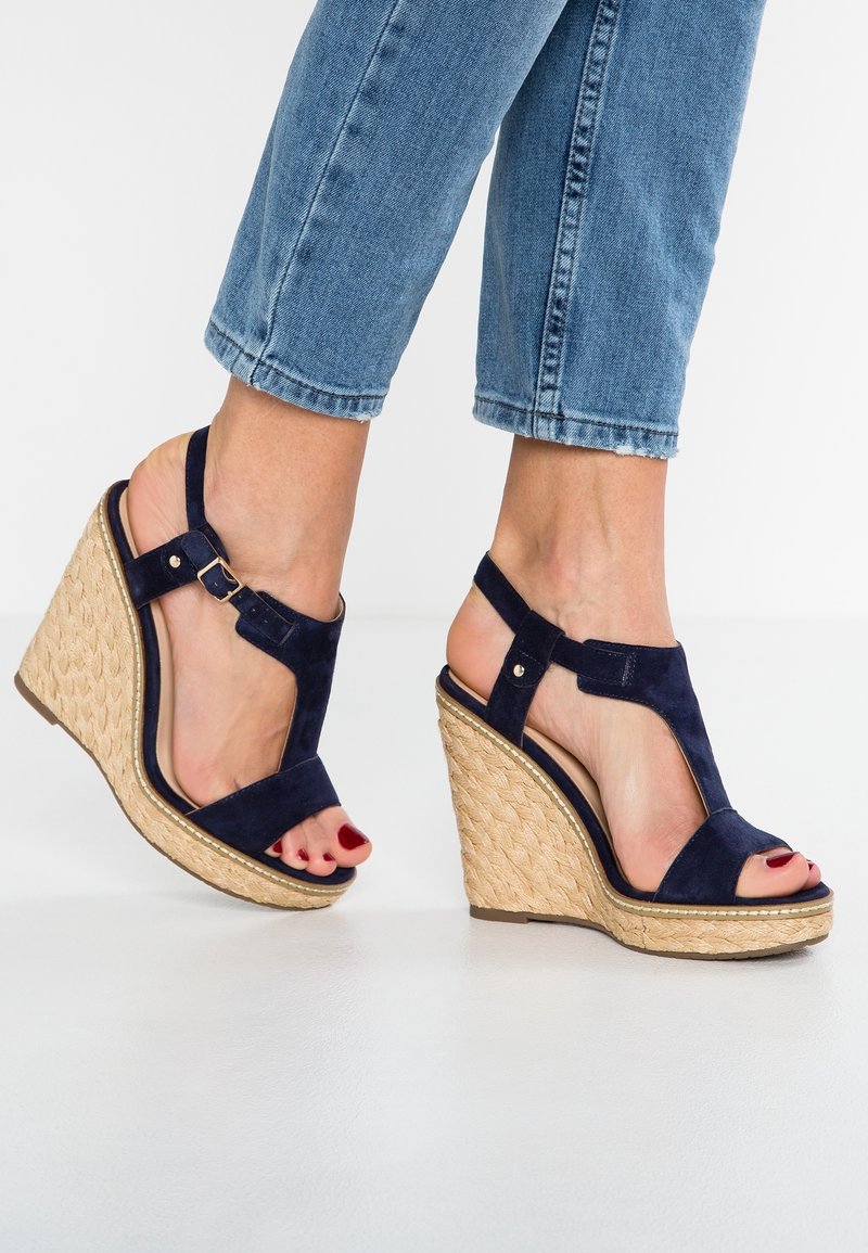 Pieds chaussés de sandales compensées en daim bleu foncé avec des lanières à la cheville et des talons plateforme tissés, associées à un jean bleu raccourci.