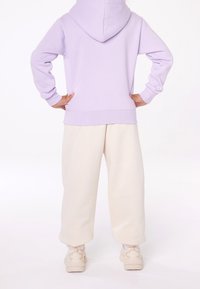 Sudadera con capucha de color lila con bolsillo canguro, combinada con pantalones cremosos de corte ajustado. La tela parece suave y texturizada. Unos zapatillas blancas completan el atuendo.