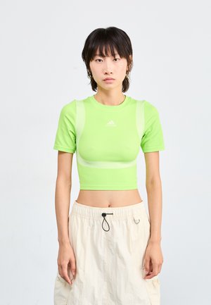 Vihreä lyhythihainen crop-top, jossa on verkkokankaita olkapäillä ja Adidaksen logo keskellä. Yhdistetty vaaleanbeigeihin elastisen vyötärön shortsit.