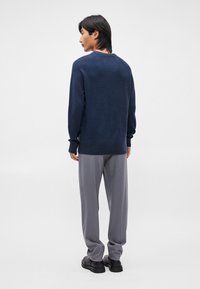 Pulover tricotat bleumarin cu manșete și tiv cu falduri, purtat cu pantaloni gri deschis și pantofi negri. Vederea din spate arată textura țesăturii.