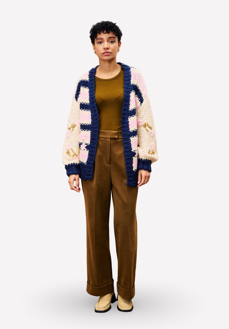 Frau mit kurzen lockigen Haaren trägt braune weit geschnittene Hose, olivgrünes Oberteil und oversized cremefarbene Strickjacke mit marinen und rosa Mustern und steht aufrecht.