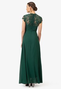 Abito da sera verde con maniche corte in pizzo, un corpino aderente e una gonna fluente. Il dettaglio in pizzo sul retro aggiunge eleganza.