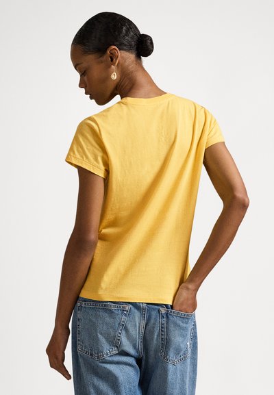 Ženska z zbito pričesko, oblečena v rumeno majico s kratkimi rokavi in modre kavbojke, obrnjena s hrbtom proti gledalcu, z eno roko v zadnjem žepu, na enakomerni podlagi.