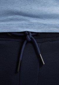Lichtblauw shirt met een zoom dat de marineblauwe sportbroek met een trekkoordsluiting ontmoet. Zwarte accenten op de uiteinden van het trekkoord; gladde stoftextuur zichtbaar.
