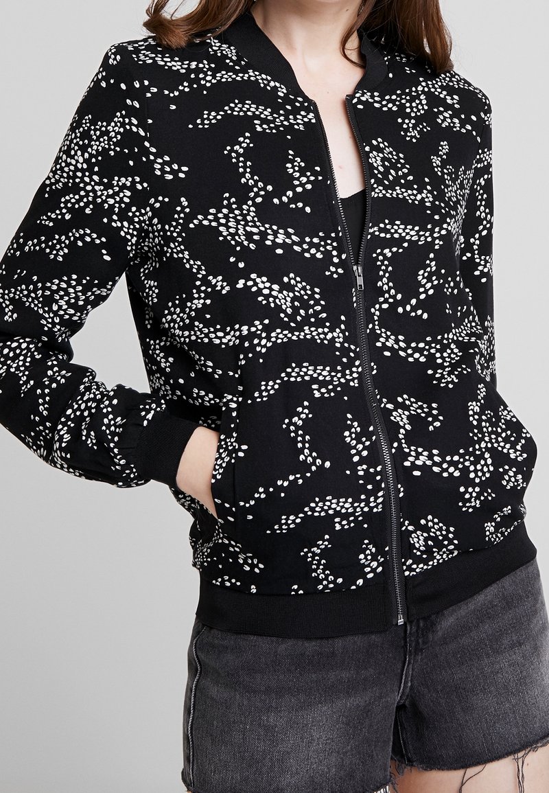 Femme portant une veste noire à zip avec des motifs abstraits de points blancs et un short en jean foncé, mains dans les poches de la veste, sur un fond uni.