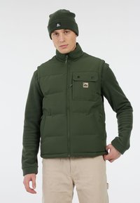 Groene puffer gilet met een rits aan de voorkant, gewatteerd ontwerp, borstzak en logo patch. Draagt over een groene fleece top, gecombineerd met beige broek.