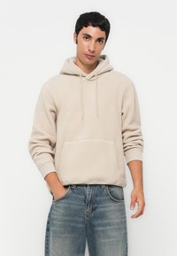Junger Mann, der einen beigen Fleece-Hoodie und blue Jeans trägt, steht mit einer Hand in der Tasche vor einem weißen Hintergrund.