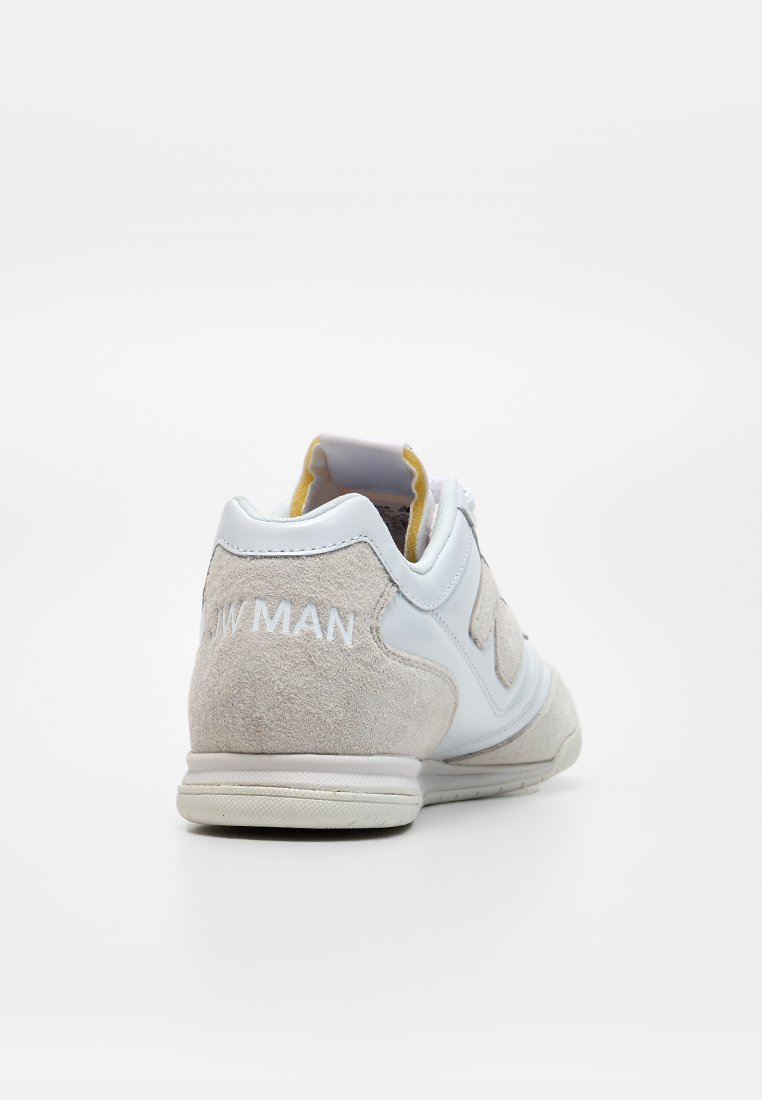 Zapatilla deportiva beige y blanca con materiales de ante y cuero, vista desde atrás, mostrando el texto "NEW MAN" en el talón.