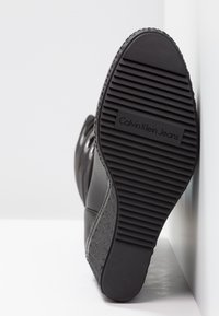 Suela de goma negra con textura acanalada, con la marca "Calvin Klein Jeans". Cuenta con una parte superior de cuero liso y un acento de tela texturizada.