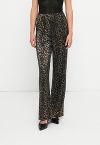 Femeie îmbrăcată în pantaloni evazați cu print leopard plisat și pantofi negri cu vârful ascuțit, stând în fața unui fundal alb simplu.