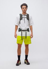 Νεαρός άνδρας που φοράει λευκό υπερμεγέθες μπλουζάκι, νεον κίτρινα σορτς, μαύρες κάλτσες, παπούτσια πεζοπορίας και ζώνη ασφαλείας με θήκες στο στήθος και τη μέση.