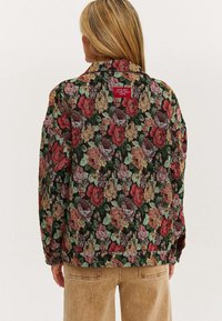 Zielona kurtka w kwiaty z czarnym tłem, pokryta teksturowanym wzorem czerwonych, różowych i zielonych róż. Posiada czerwoną metkę z napisem z tyłu. Luźny krój.