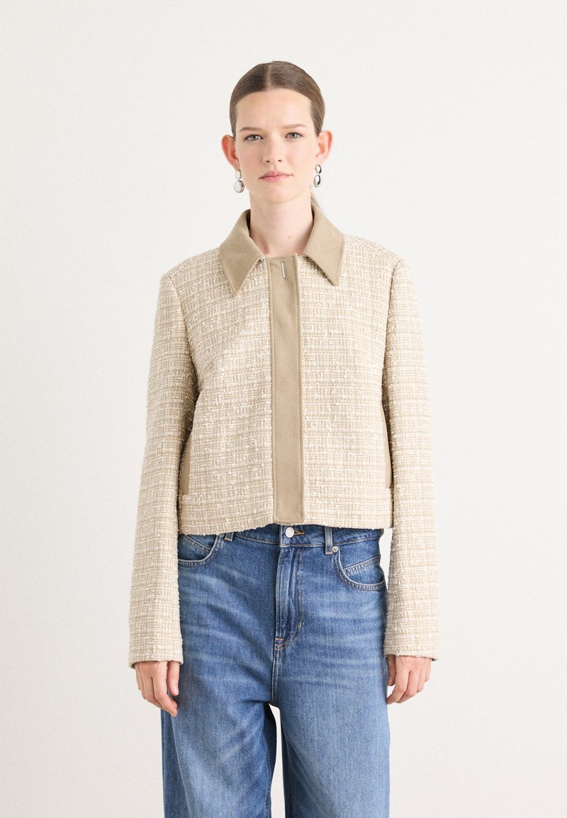 Beige knappe Jacke mit strukturiertem Gewebe und schmalem Kragen. Mit einem Reißverschluss vorne, langen Ärmeln und einer kastigen Silhouette. Getragen mit blauen Jeans.