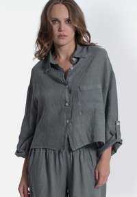 Camicia button-up in lino grigia con una vestibilità ampia, maniche lunghe e taschino frontale. Presenta dettagli con bottoni chiari e polsini delle maniche arrotolati.