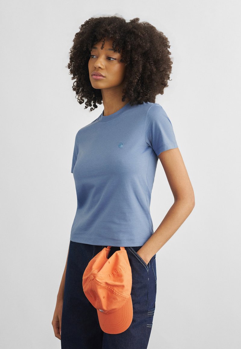 Frau mit lockigem Haar, die ein eng anliegendes blaues T-Shirt und dunkle Jeans trägt und eine orangefarbene Baseballkappe in der Hand hält.