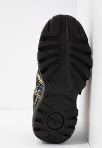 Svart gummisula för sneakers med texturerat greppmönster. Gula och vita accenter längs sidorna och en distinkt design på ovansidan.