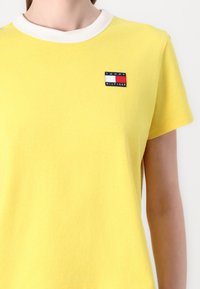 Gul bomulds t-shirt med en hvid ribkant i kraven og et lille broderet logo på brystet med et rektangulært design i rød, hvid og blå.