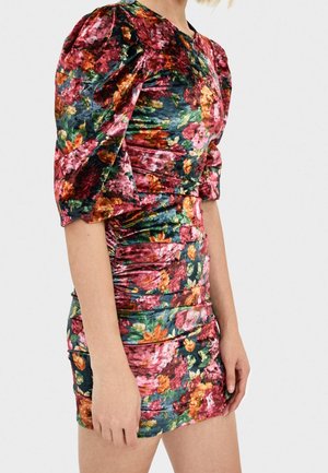 Kobieta ubrana w dopasowaną, marszczoną aksamitną sukienkę z kolorowym kwiatowym wzorem i bufiastymi krótkimi rękawami, stojąca na jednolitym tle.